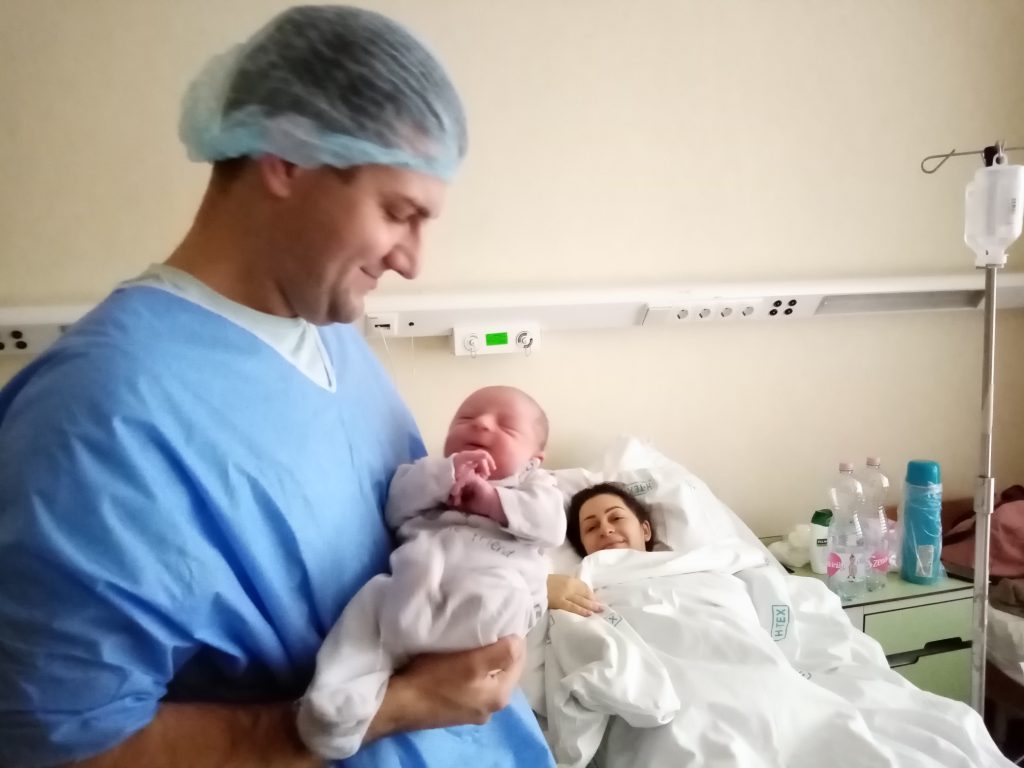 Édesapja karjaiban az újszülött Kornél Márton, háttérben az édesanya pihen a kórház szülészetén található családi szobában