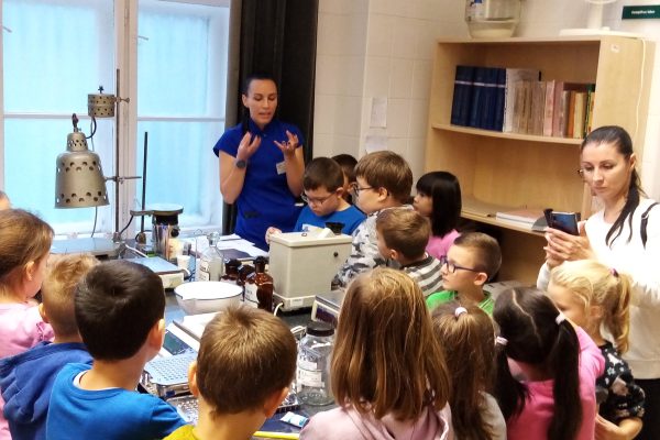 a képen a gyerekek a gyógyszerésztár laboratóriumában körbeállják a laborasztalt és figyelik, ahogy a gyógyszerész a szükséges alapanyagokat kiméri, majd a gyerekek saját kézzel keverik ki a levendulás kézkrémet