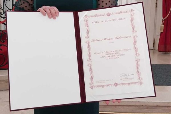 A fotón Balázsné Mészáros Ildikó látható, a fővárosi Stefánia palotában, a kezében van a miniszteri elismerő oklevél