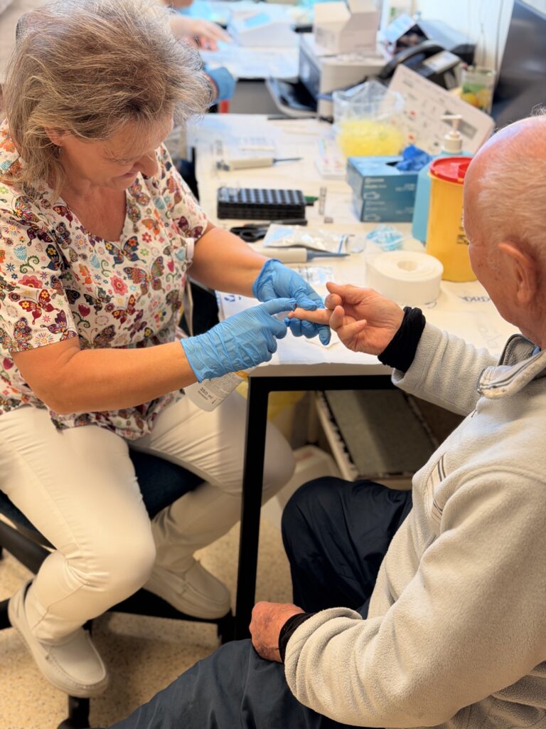 Egészségügyi dolgozó gumikesztyűben ujjbegyből vért vesz egy idős férfitól asztal mellett ülve.