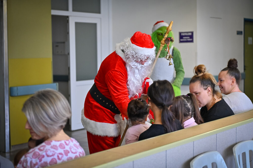 A képen egy kórházi folyosón vagy váróteremben zajló ünnepi jelenet látható. Középen egy Mikulás-ruhás férfi áll, fehér szakállal és piros sapkában, kezében hosszú bottal, amelyen kis csengők vannak. A Mikulás több, padon ülő gyermek felé hajol, és ajándékot ad át nekik. A gyerekeket háttal ülve felnőttek tartják. A háttérben egy zöld jelmezbe öltözött figura – a Grincs – áll, szintén ünnepi kiegészítővel.