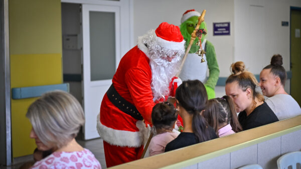 A képen egy kórházi folyosón vagy váróteremben zajló ünnepi jelenet látható. Középen egy Mikulás-ruhás férfi áll, fehér szakállal és piros sapkában, kezében hosszú bottal, amelyen kis csengők vannak. A Mikulás több, padon ülő gyermek felé hajol, és ajándékot ad át nekik. A gyerekeket háttal ülve felnőttek tartják. A háttérben egy zöld jelmezbe öltözött figura – a Grincs – áll, szintén ünnepi kiegészítővel.