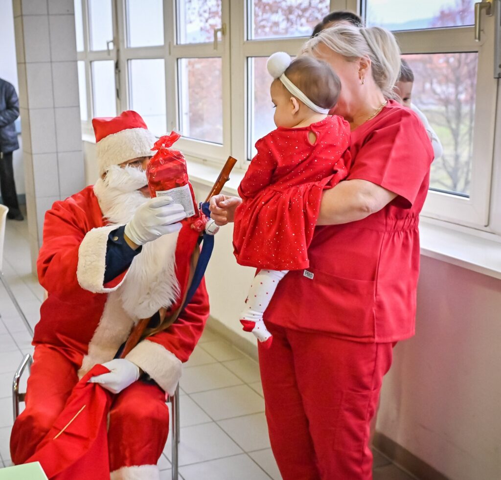 kórházi folyosón Mikulás ruhában személy ül egy széken. Fehér szakállat, piros sapkát, piros kabátot és fehér kesztyűt visel. A kezében egy piros csomagolású ajándék csomagot tart, amelyet egy kisbaba felé nyújt, akit egy piros ruhás egészségügyi dolgozó tart.