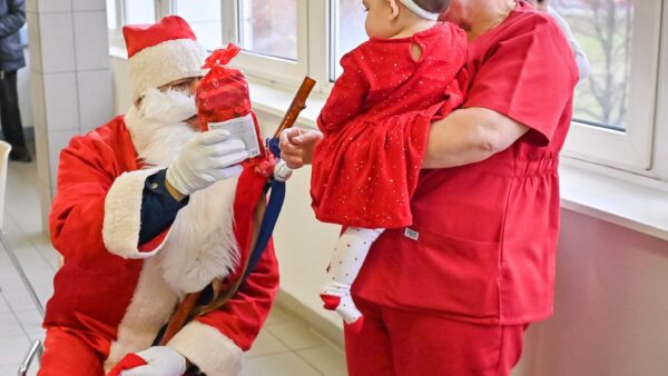 kórházi folyosón Mikulás ruhában személy ül egy széken. Fehér szakállat, piros sapkát, piros kabátot és fehér kesztyűt visel. A kezében egy piros csomagolású ajándék csomagot tart, amelyet egy kisbaba felé nyújt, akit egy piros ruhás egészségügyi dolgozó tart.