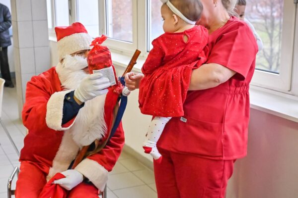 kórházi folyosón Mikulás ruhában személy ül egy széken. Fehér szakállat, piros sapkát, piros kabátot és fehér kesztyűt visel. A kezében egy piros csomagolású ajándék csomagot tart, amelyet egy kisbaba felé nyújt, akit egy piros ruhás egészségügyi dolgozó tart.
