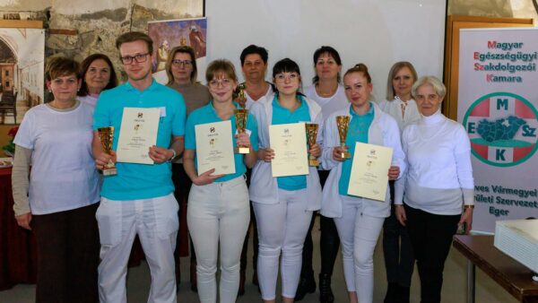 A képen a XIII. „Hivatásunk mesterei vagyunk” szakdolgozói verseny díjazott csapata látható az oklevelekkel és kupákkal a kezükben, a szervezők és kollégák körében. A résztvevők ünnepélyes keretek között, a Magyar Egészségügyi Szakdolgozói Kamara molinója előtt állva örökítették meg a közös sikert.