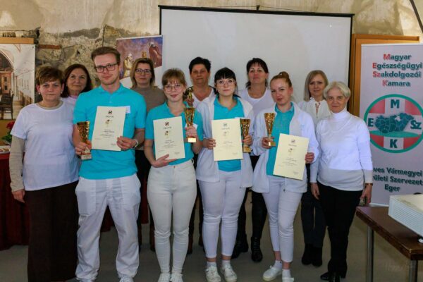 A képen a XIII. „Hivatásunk mesterei vagyunk” szakdolgozói verseny díjazott csapata látható az oklevelekkel és kupákkal a kezükben, a szervezők és kollégák körében. A résztvevők ünnepélyes keretek között, a Magyar Egészségügyi Szakdolgozói Kamara molinója előtt állva örökítették meg a közös sikert.