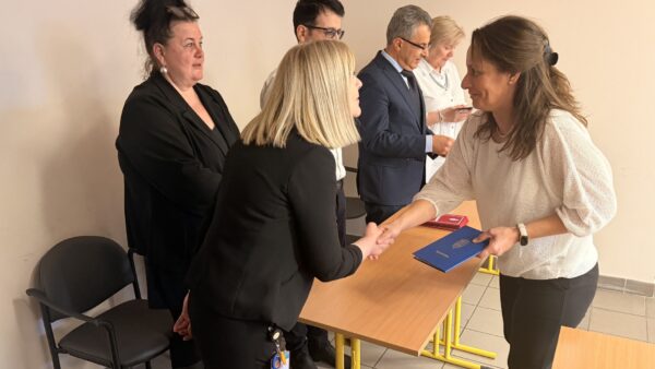 Az ünnepségen az egyik végzős hallgató átveszi bizonyítványát, miközben kézfogással gratulálnak neki az átadó asztalnál álló vezetők és oktatók.