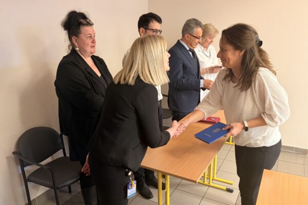 Az ünnepségen az egyik végzős hallgató átveszi bizonyítványát, miközben kézfogással gratulálnak neki az átadó asztalnál álló vezetők és oktatók.