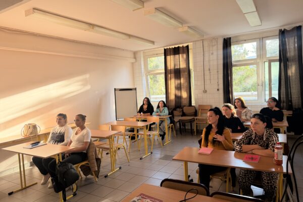 A „Nő az esély” program résztvevői egy világos tanteremben, egymás mögött elhelyezett asztaloknál ülnek. A teremben világos színű, sárga lábú székek, nagy ablakok és fehér falak láthatók.
